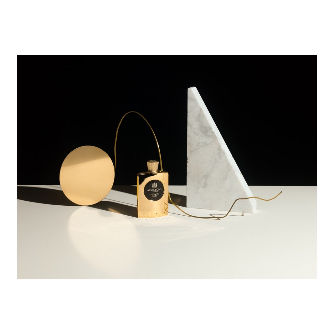 A gold perfume bottle takes center stage in a minimalist still life, complemented by a gold disc and a white marble triangle.