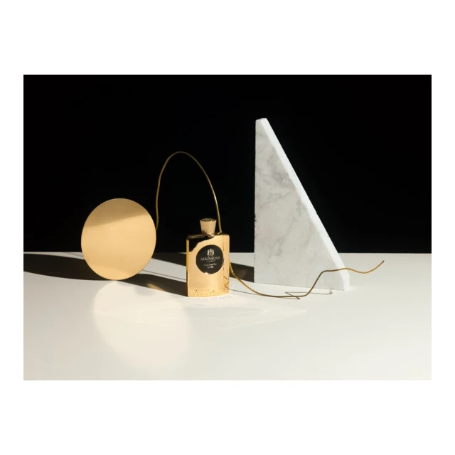 A gold perfume bottle takes center stage in a minimalist still life, complemented by a gold disc and a white marble triangle.
