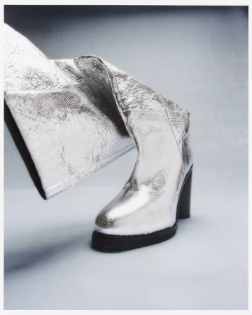 Metallic silver boot with platform sole and block heel, paired with matching silver pants. Fashionable footwear on a light backdrop.