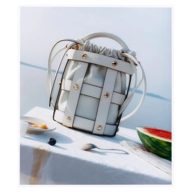 Stylish white woven handbag with gold accents displayed next to fruit on a white surface with soft focus background. Fashion still life.