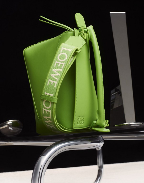 Bright green Loewe bucket bag with branded strap sits on a reflective surface against a dark background in a studio product shot.