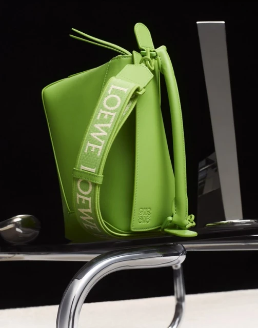 Bright green Loewe bucket bag with branded strap sits on a reflective surface against a dark background in a studio product shot.