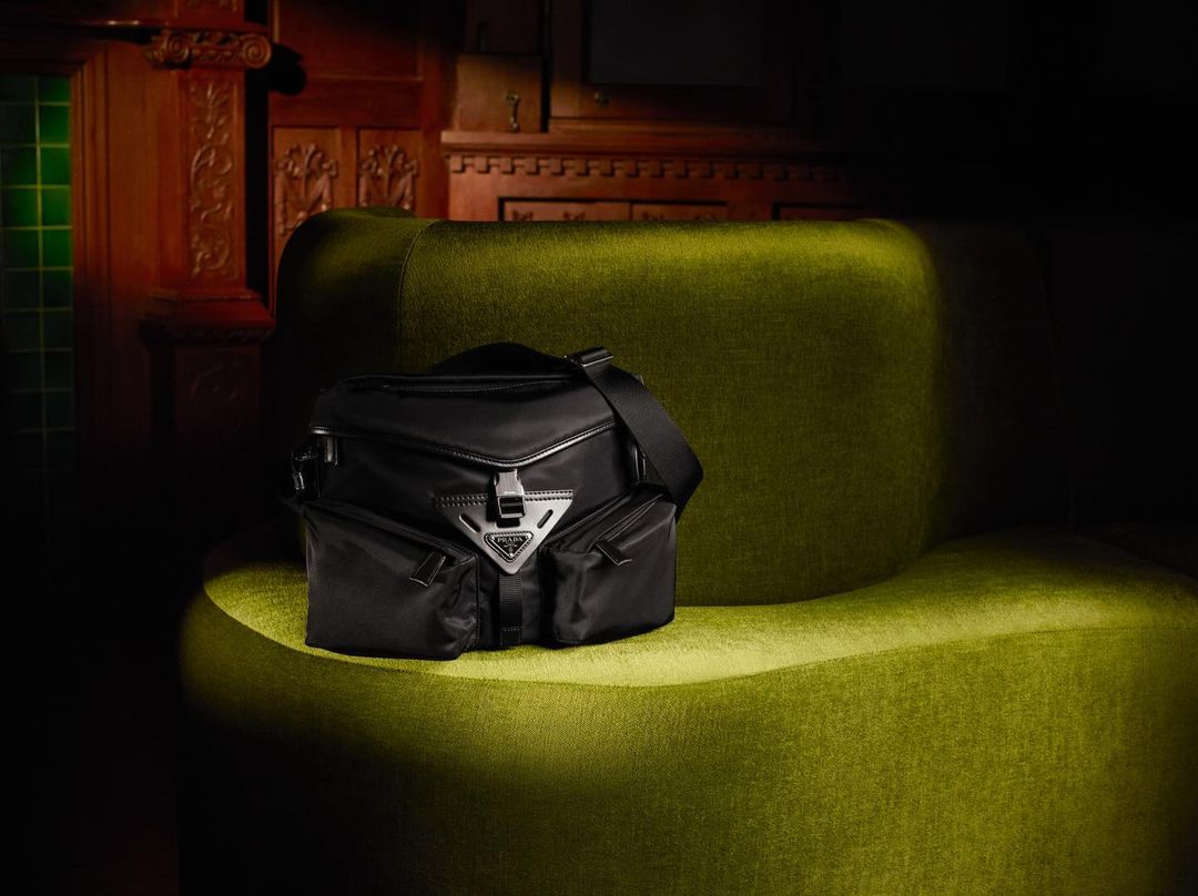 Black Prada handbag with silver hardware and logo placed on an olive green velvet chair against a dark wood paneled wall.