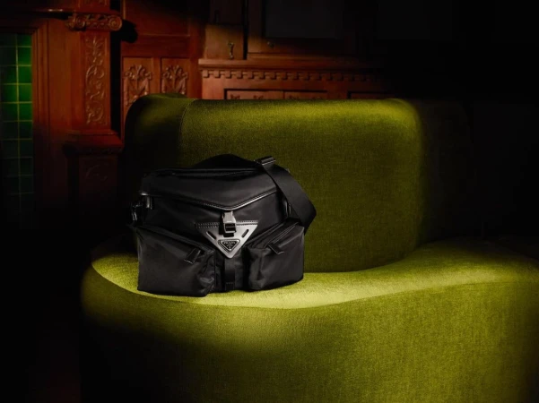Black Prada handbag with silver hardware and logo placed on an olive green velvet chair against a dark wood paneled wall.