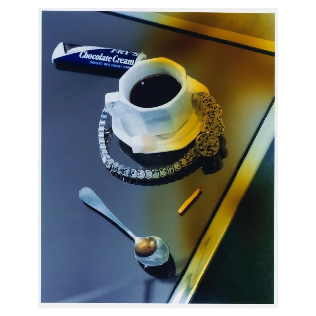 Elevated view of a cup of coffee next to Fry's chocolate bar, a silver spoon, gold coins, and a bracelet on a reflective dark surface.