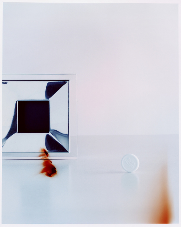 Close-up still life photograph featuring a small white round tablet and a metallic square object on a white surface.