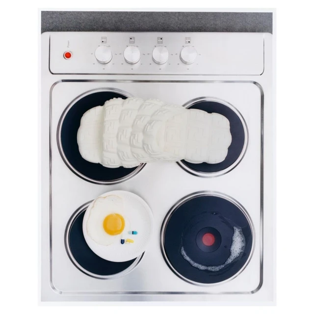 Overhead shot of an electric stove with a designer shoe and a plate with a fried egg and pills on it, creating a surreal still life.