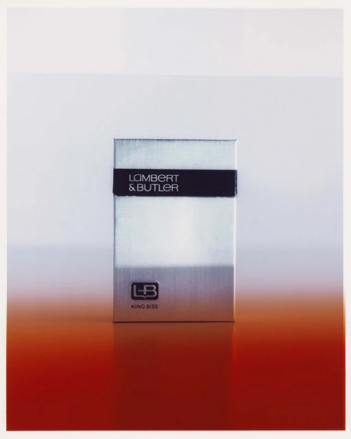 Close-up studio shot of a silver Lambert & Butler cigarette box, with a red gradient at the bottom, against a gradient white backdrop.