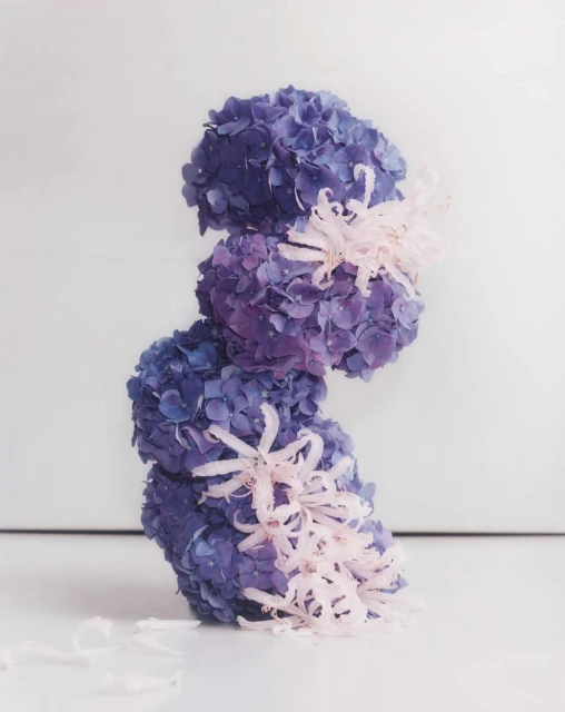 Still life of three stacked purple hydrangeas with white decorative accents against a clean, bright white backdrop and surface.