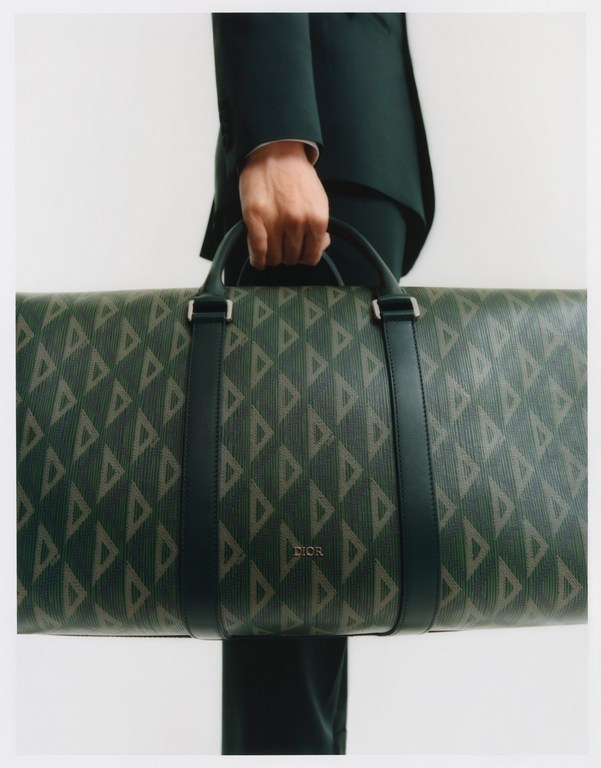 Close-up of a person holding a large green and beige Dior travel bag with a geometric pattern against a neutral background in a studio.