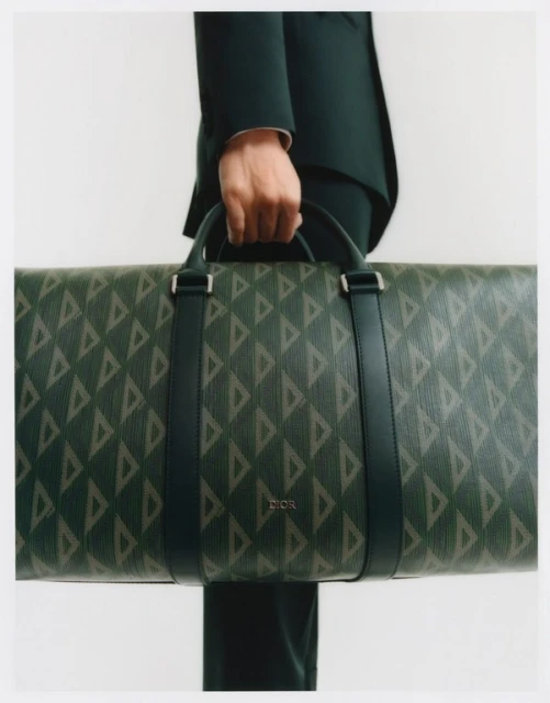Close-up of a person holding a large green and beige Dior travel bag with a geometric pattern against a neutral background in a studio.