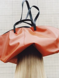 Close-up of a person wearing a large orange handbag balanced on their head with long straight blonde hair visible below