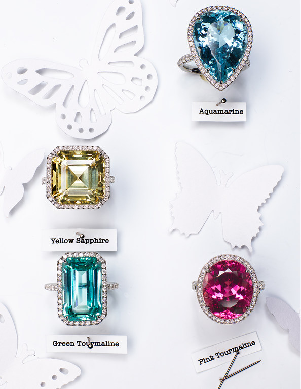 Four rings with different colored gems against a white background decorated with white butterfly paper cutouts in a bright studio shot.