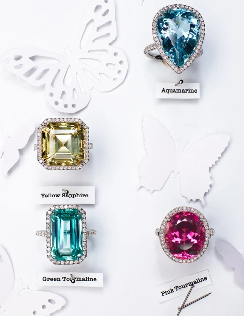 Four rings with different colored gems against a white background decorated with white butterfly paper cutouts in a bright studio shot.