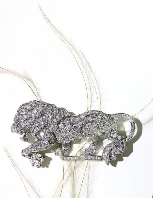 Close-up of a decorative silver brooch shaped like a lion encrusted with gems, set against a backdrop of delicate white feathers.