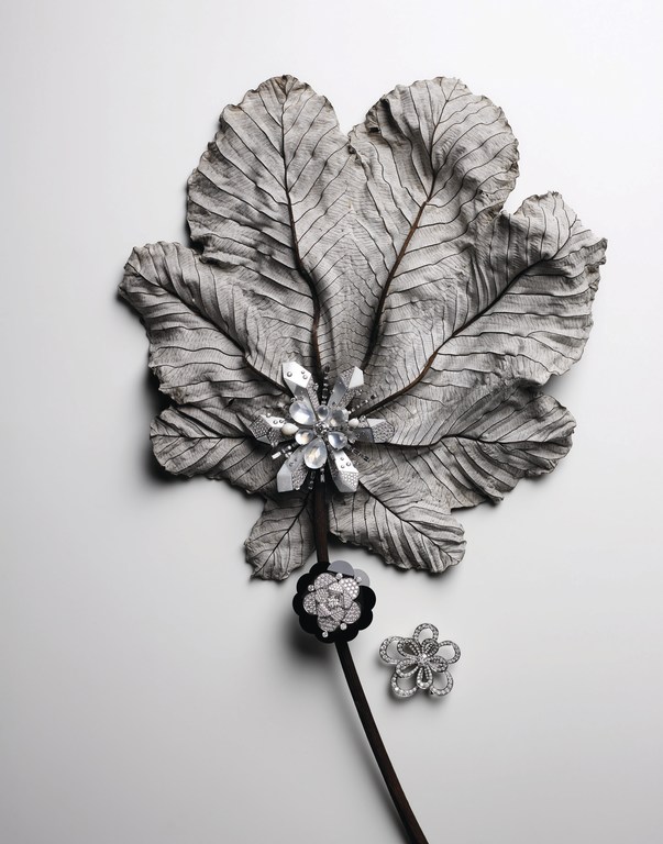 Still life featuring a large, textured grey leaf with decorative flower brooches in silver and black on a white background.