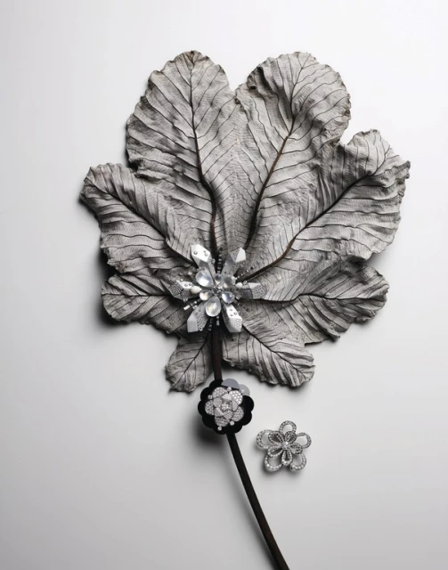 Still life featuring a large, textured grey leaf with decorative flower brooches in silver and black on a white background.