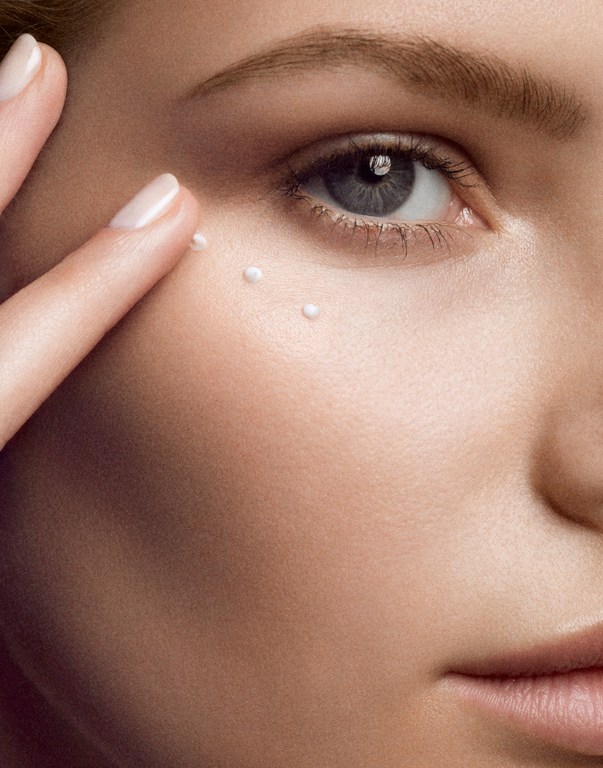 Close-up of a woman applying skincare cream under her eye with her finger, showcasing beauty and skincare in a neutral setting.
