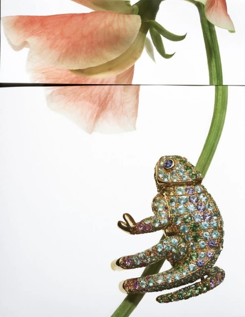 Chameleon-shaped jeweled object climbs a thin green stalk next to a pale pink flower against a white background in a studio shot.
