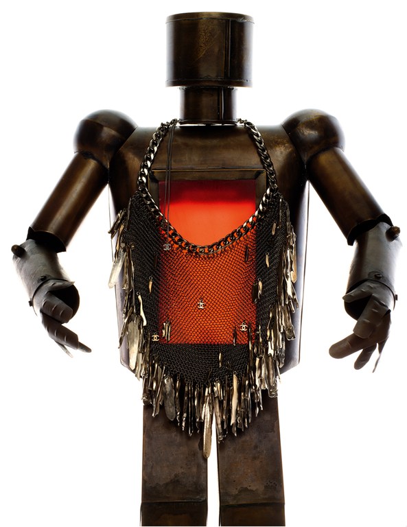 Metal robot figure with a metallic mesh garment that has tear-drop shaped metal pieces attached to the bottom, against a white backdrop.