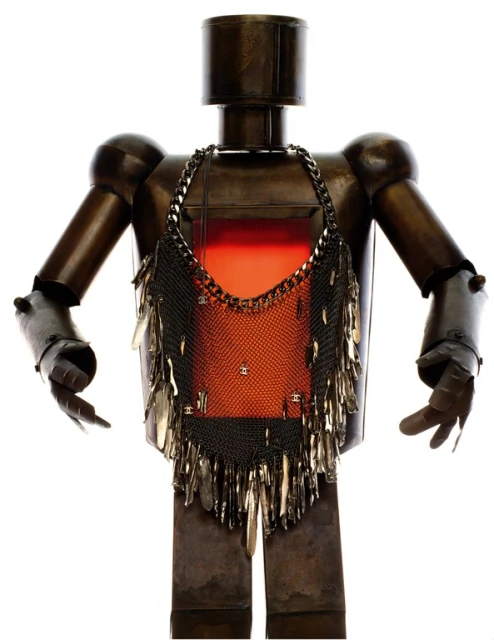 Metal robot figure with a metallic mesh garment that has tear-drop shaped metal pieces attached to the bottom, against a white backdrop.