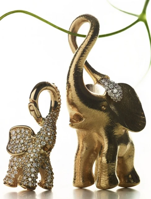 Two golden elephant pendants with diamond embellishments are displayed in a studio setting with a green vine accent.