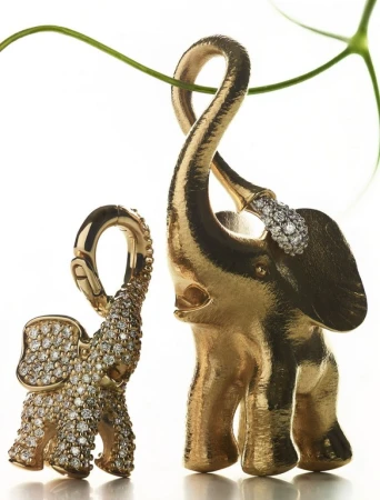 Two golden elephant pendants with diamond embellishments are displayed in a studio setting with a green vine accent.