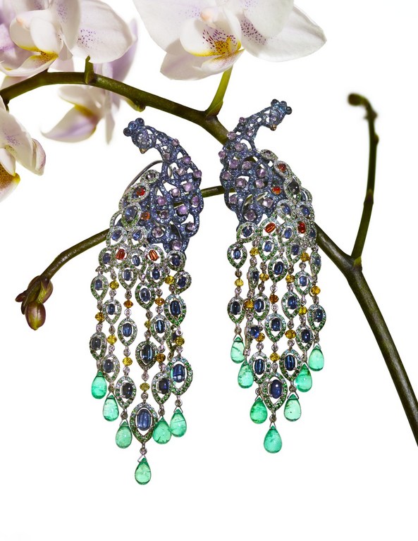 Close-up of two ornate peacock-shaped dangling earrings with colorful gemstones, hanging on a flowering branch against a white background.