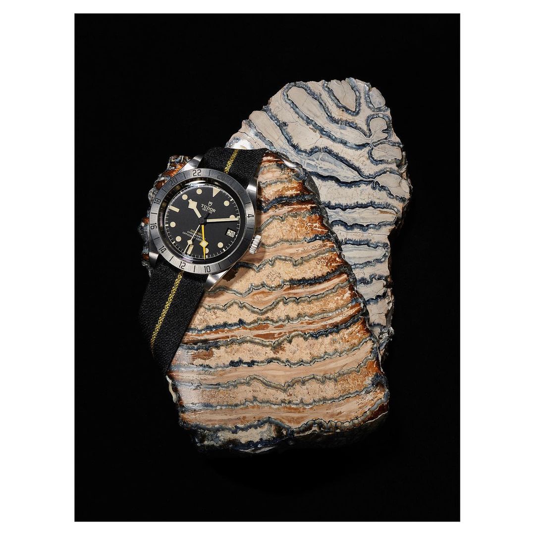 Luxury watch on a striped fabric band presented on a layered rock slab against a black background in a studio-style product shot.