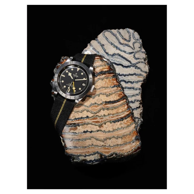 Luxury watch on a striped fabric band presented on a layered rock slab against a black background in a studio-style product shot.