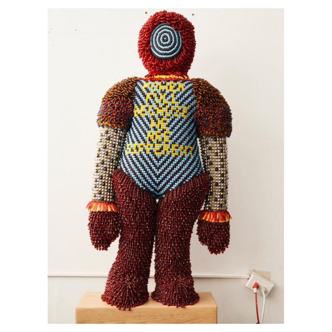 Sculptural beaded figure standing on a wooden base against a white wall, featuring textured beadwork in red, blue, and metallic colors.