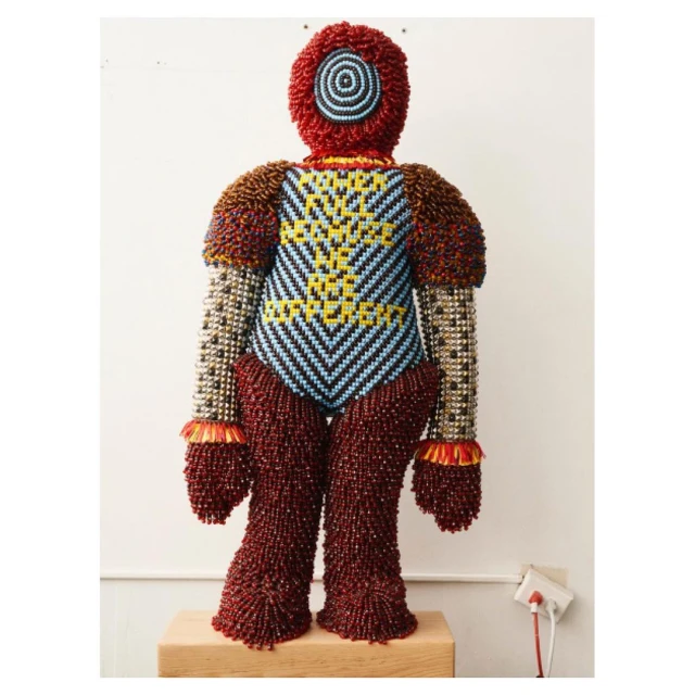 Sculptural beaded figure standing on a wooden base against a white wall, featuring textured beadwork in red, blue, and metallic colors.