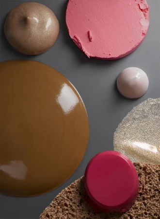 Overhead shot of various makeup textures and colors, including foundation, blush, and highlighter on a grey surface with sponge.