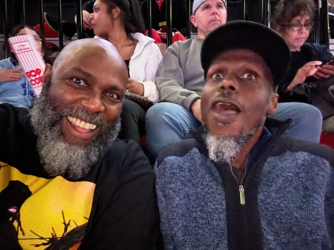 Two adult men pose for a selfie at a sporting event, one smiling broadly and the other looking surprised, with stadium seating visible in the background.
