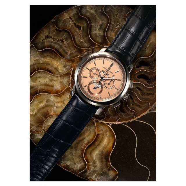 Luxury wristwatch with a brown leather strap and silver casing displayed on a patterned stone slab against a dark background.