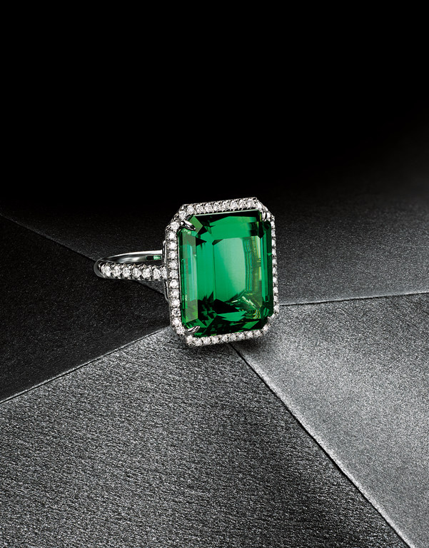 Close-up of an emerald-cut green gemstone ring with diamond accents on a dark grey textured surface, spotlighting jewelry design.