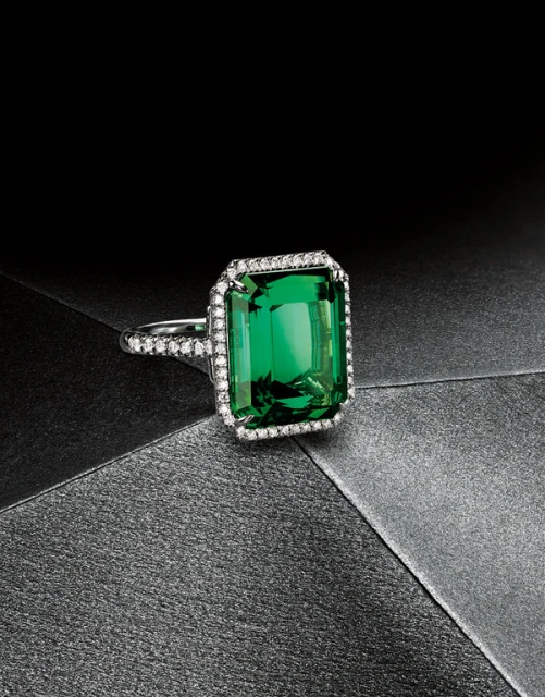 Close-up of an emerald-cut green gemstone ring with diamond accents on a dark grey textured surface, spotlighting jewelry design.