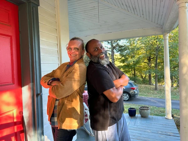 A smiling man and woman standing back-to-back with arms crossed on a covered porch with a bright red door.