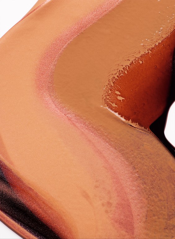 Close-up of swatches of different shades of brown and tan foundation or concealer makeup on a white surface.