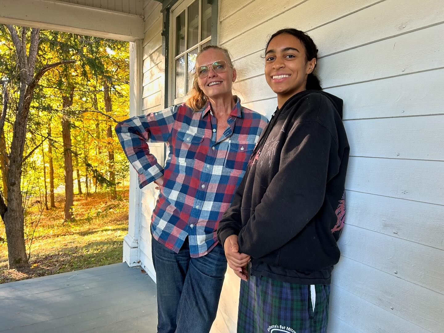 Two women pose on a porch with an autumnal wooded backdrop, one in a red and blue plaid shirt and the other in a black hoodie.