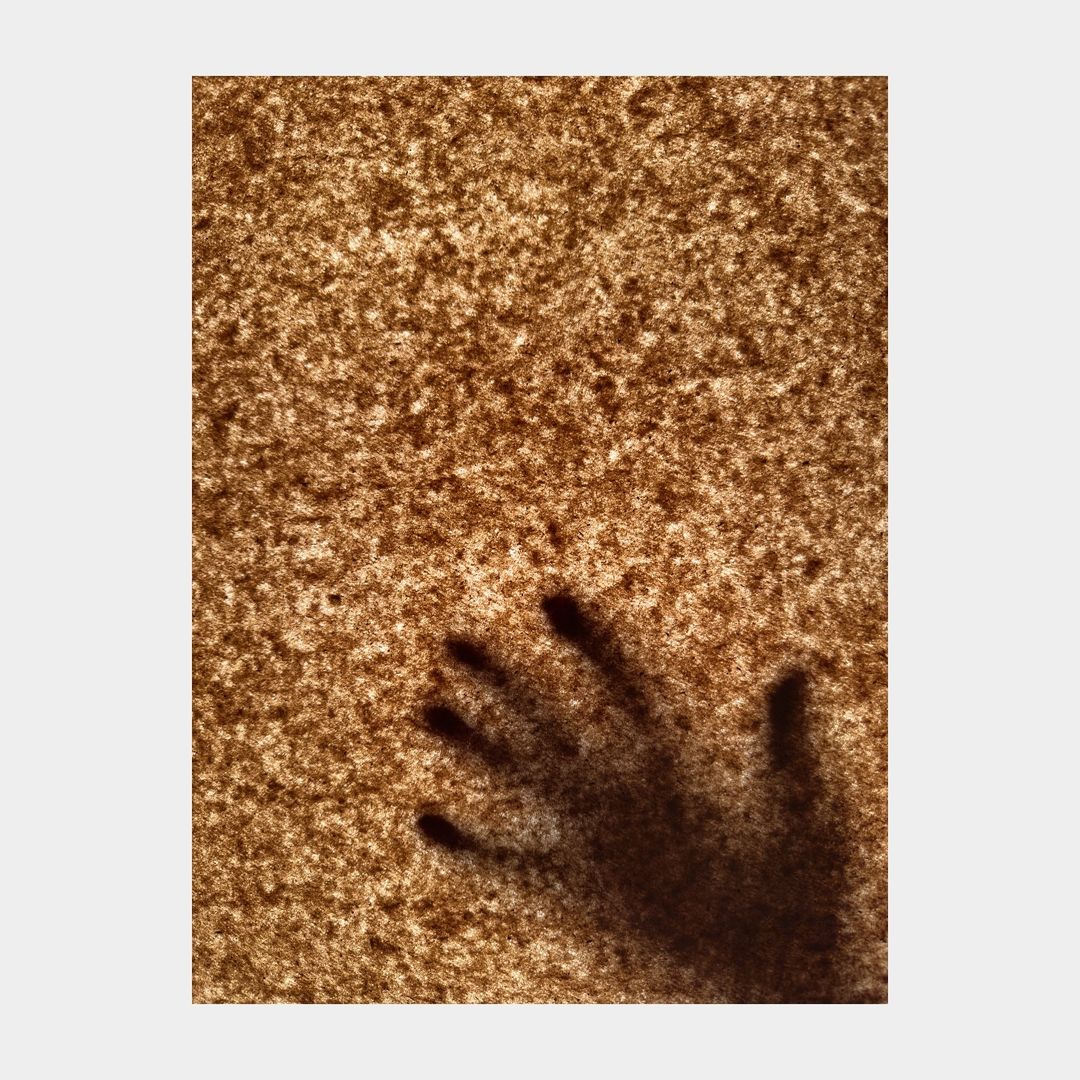 Shadow of a hand with fingers spread against a textured, mottled brown background, creating an eerie, abstract, and somewhat ominous effect.