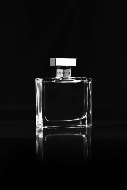 Monochromatic studio shot of a clear, square perfume bottle with a silver cap on a reflective surface against a dark background.