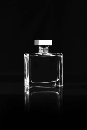 Monochromatic studio shot of a clear, square perfume bottle with a silver cap on a reflective surface against a dark background.