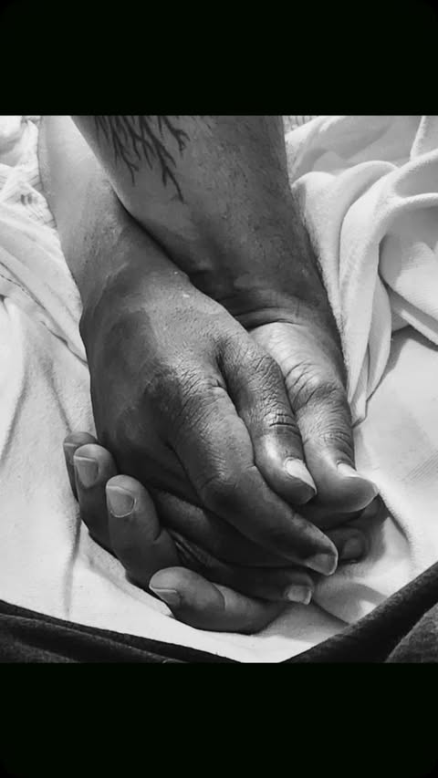 Close-up, black and white image of two hands clasped together on a white surface with a tattoo visible on one of the wrists.