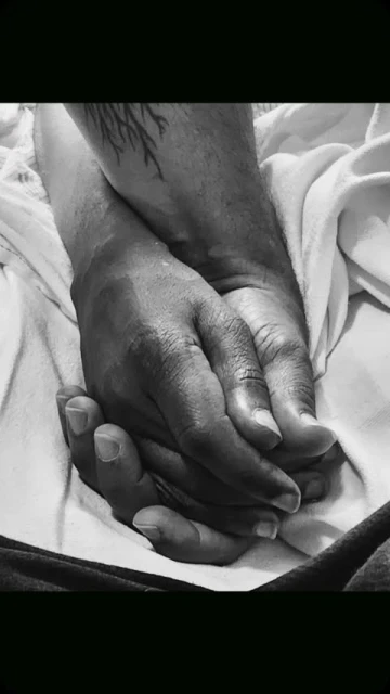 Close-up, black and white image of two hands clasped together on a white surface with a tattoo visible on one of the wrists.
