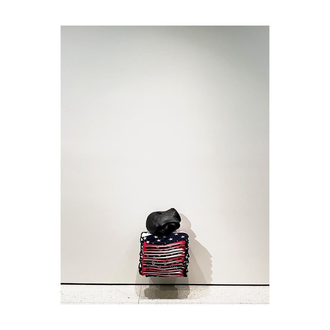 American flag artwork, folded and stacked with a black soft sculpture on top, set against a minimalist light gray interior.