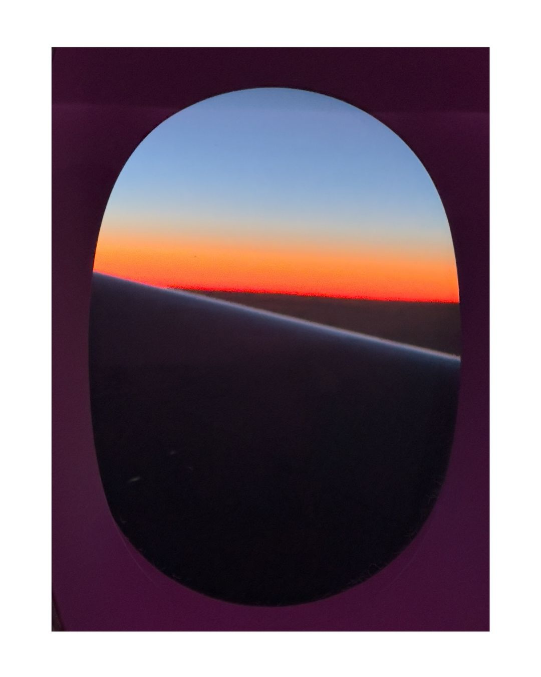 Airplane window view of colorful sunset sky with orange and blue hues. Airplane wing visible through window.