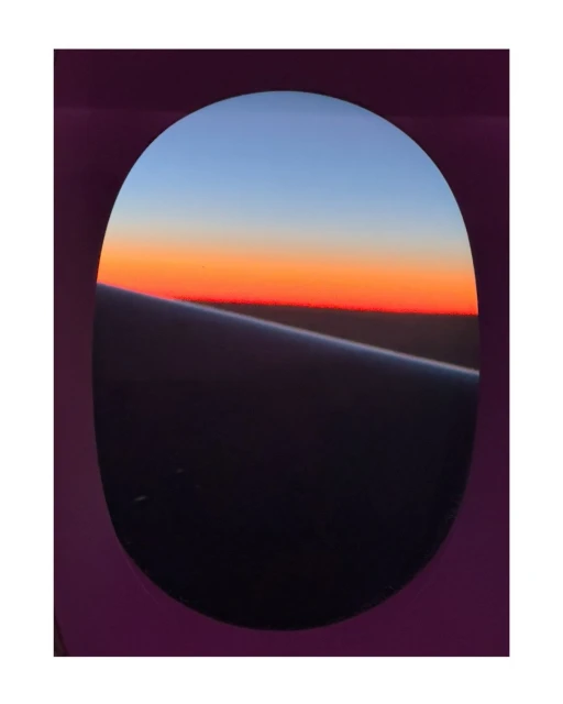 Airplane window view of colorful sunset sky with orange and blue hues. Airplane wing visible through window.
