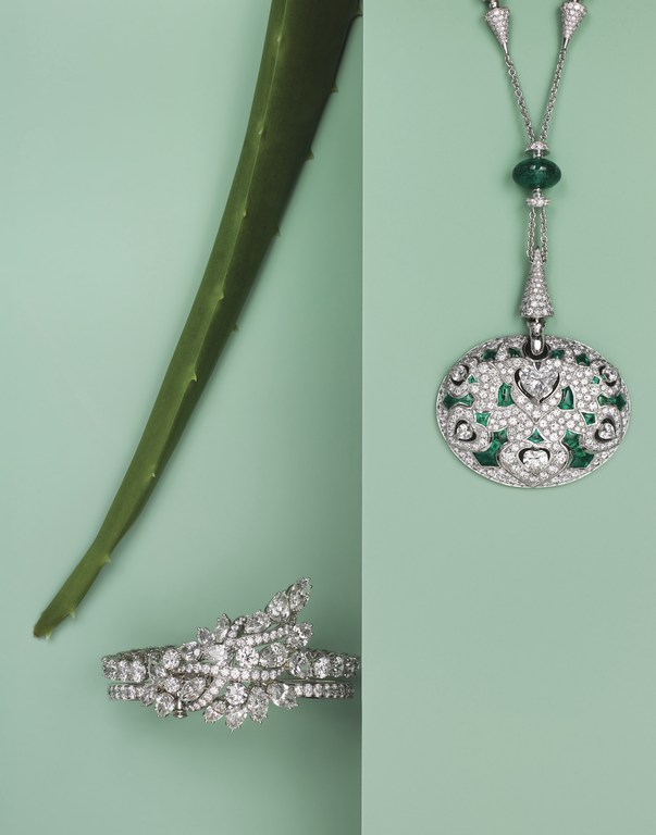 An overhead shot showcases luxury jewelry: a necklace and bracelet featuring diamonds and emeralds, alongside an aloe vera leaf on a light green surface.