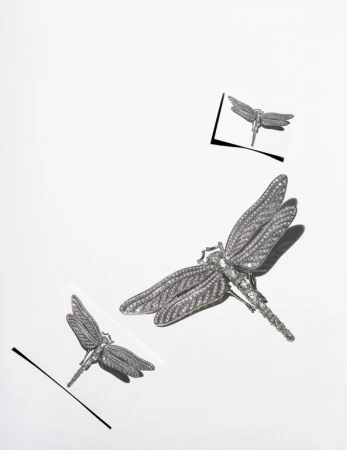 Three silver dragonfly brooches are displayed on a white surface with two of the brooches also displayed on clear squares.